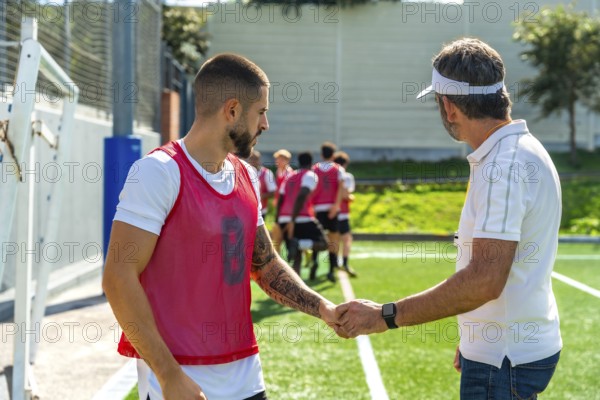Coach and young soccer player shaking hands on a green sports field, demonstrating teamwork, respect, and sportsmanship during training with other teammates in the background