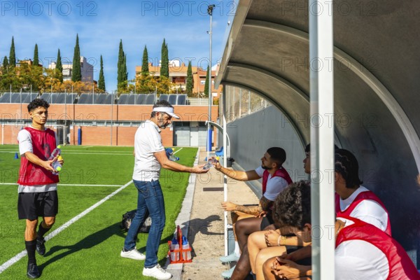 Soccer coach distributing water bottles to young male players sitting in the dugout during a hydration break on the field after an intensive training session