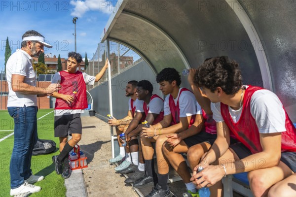 Soccer coach talking and giving instructions to his diverse team of young male athletes sitting on the bench during a training session or match, focusing on strategy and teamwork