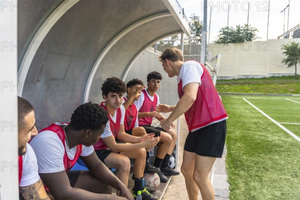 Youth soccer coach discussing strategy with his multiethnic male team members on the bench of a sports field during a training session or game, emphasizing teamwork and guidance