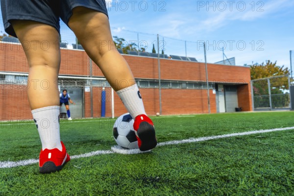 Woman soccer player preparing to strike a penalty kick on a green artificial turf field, with a blurred goalkeeper in the background ready to defend the goal