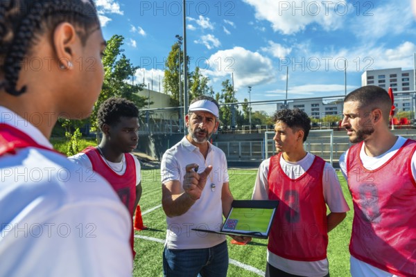 Soccer coach explaining game strategy to a diverse group of male players on a green artificial turf field, showing teamwork, coaching, and a professional plan during training