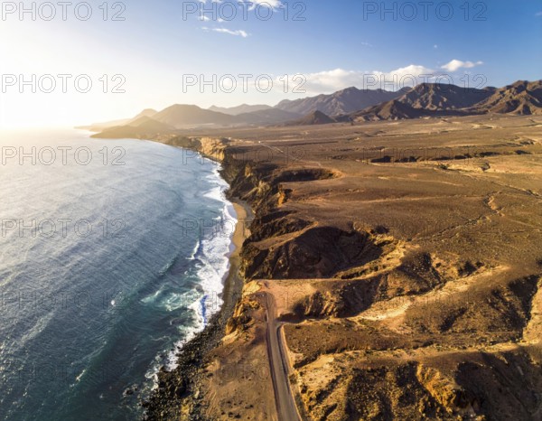 Desert landscape converges with ocean along a rugged coastal area, shore break at cliffs, blue cloudy sky at sunset, dry landscape in an aerial drone view, AI generated