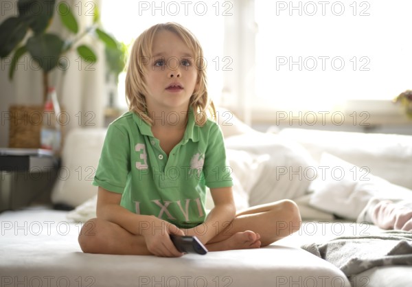 Child, boy, 7 years old, blonde, sitting on sofa, with remote control in hand, watching TV, watching television, watching television, excited, fascinated, Stuttgart, Baden-Württemberg, Germany