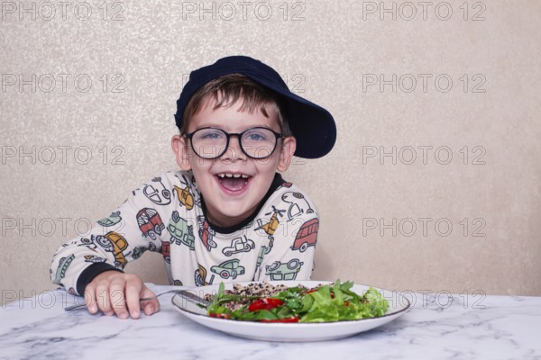 Child reacts dramatically to a salad served on a marble table at home during lunchtime, A little boy in glasses, and a cap, makes a funny face, sitting at a marble table. A salad is placed in front of him, demonstrating, his humorous reaction, during lunch