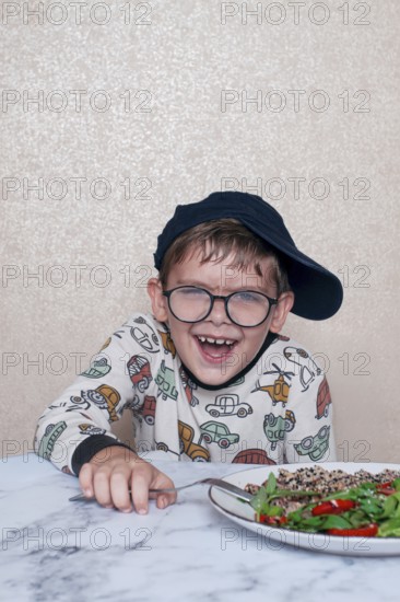 Child reacts dramatically to a salad served on a marble table at home during lunchtime, A little boy in glasses, and a cap, makes a funny face, sitting at a marble table. A salad is placed in front of him, demonstrating, his humorous reaction, during lunch