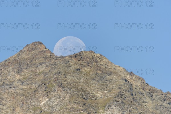 Majestic mountain peak stands tall against vibrant blue sky as large moon rises just behind it. The scene captures the quiet beauty of nature in the early evening light. Grachen, Viege, Valais, Switzerland