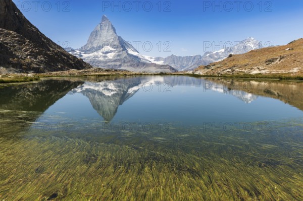 The sharp peak of Matterhorn rises majestically above a serene alpine lake, reflecting perfectly on the still water. The lush green of the grass lines the shoreline in this beautiful landscape. Zermatt, Valais, Alps, Swiss
