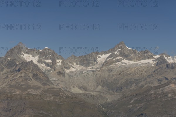 Majestic mountain peak rises prominently against a vibrant blue sky, surrounded by rocky terrain and snow-covered slopes. The sun illuminates the landscape as the clouds drift nearby. Zermatt, Valais, Alps, Swiss