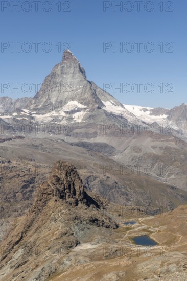 The towering peak of the Matterhorn dominates the clear sky, surrounded by rocky terrain and patches of snow. The barren landscape reveals the natural beauty of the Swiss Alps. Zermatt, Valais