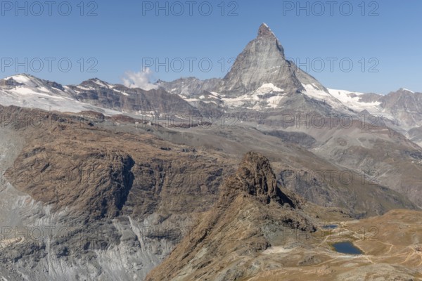 The towering peak of the Matterhorn dominates the clear sky, surrounded by rocky terrain and patches of snow. The barren landscape reveals the natural beauty of the Swiss Alps. Zermatt, Valais