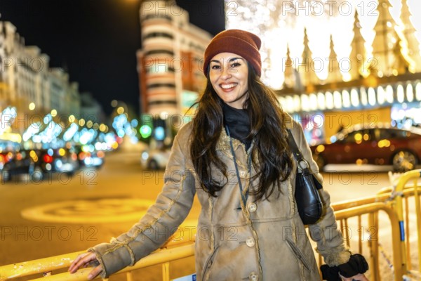 Young woman wearing winter clothes smiling in a city street decorated with christmas lights at night