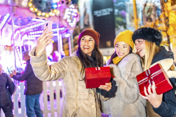 Three happy women holding christmas gifts are taking a selfie in front of a carousel in a christmas market at night
