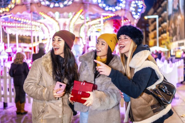 Three happy female tourists holding christmas gifts pointing at something at the christmas market carousel by night