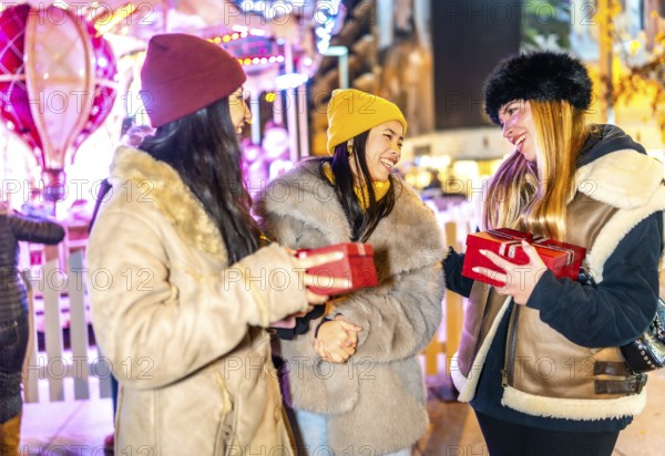 Three cheerful female friends exchanging christmas gifts at a night market with illuminated carousel in background