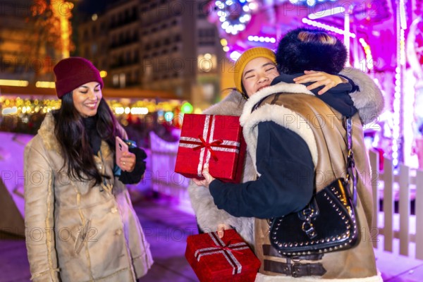 Three happy female friends exchanging christmas gifts and hugging at a christmas market at night