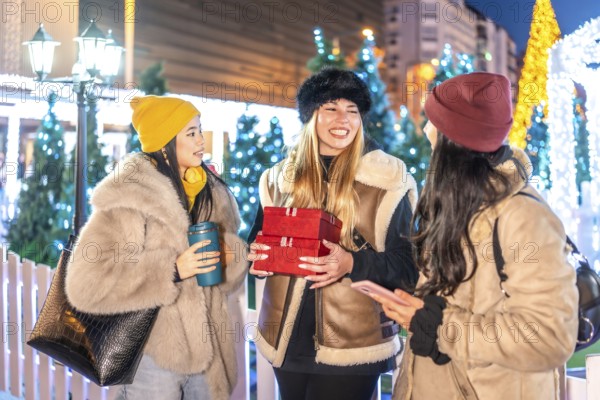 Three cheerful young women holding christmas gifts and hot drinks, enjoying a winter evening in a festively decorated city