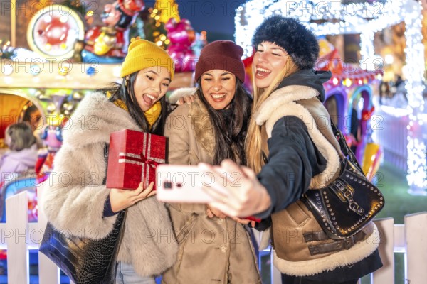 Three happy female friends taking a selfie with a smartphone at a christmas market amusement park, holding christmas gifts