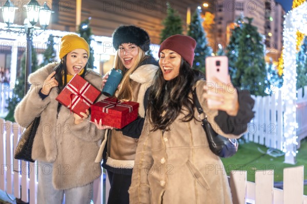 Three cheerful female friends holding christmas gifts and taking a selfie at a decorated outdoor christmas market at night