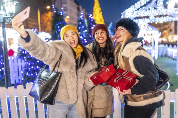 Three cheerful women taking selfie with christmas gifts and sticking out tongue in christmas market at night