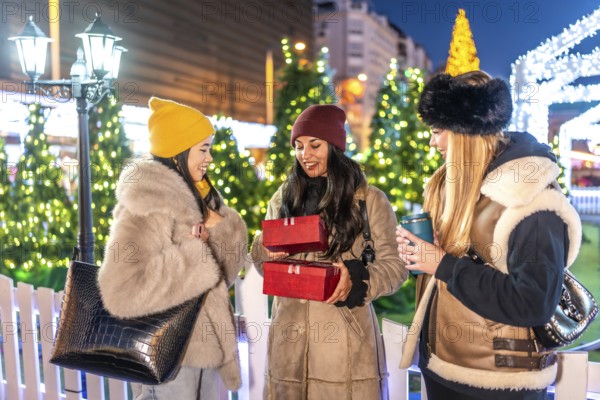 Three friends exchanging christmas presents and enjoying hot drinks at a decorated outdoor christmas market in a city at night