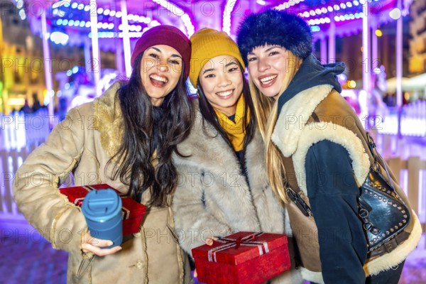 Three happy female friends enjoying christmas market at night, holding gifts and hot beverages, with illuminated carousel in background