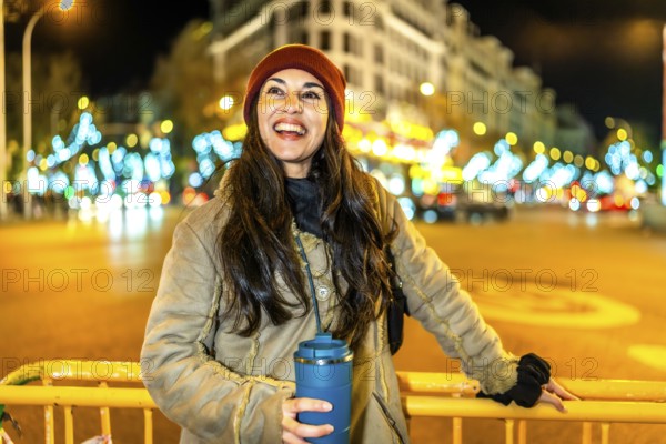 Young woman holding reusable mug smiling near christmas lights decorations in a city at night