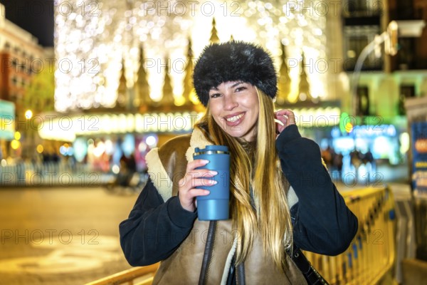 Smiling woman with winter clothes and hot beverage enjoying christmas lights and attractions in a city at night