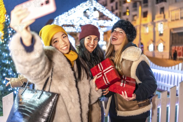 Three cheerful women holding christmas gifts taking a selfie with smartphone at christmas market in the city at night