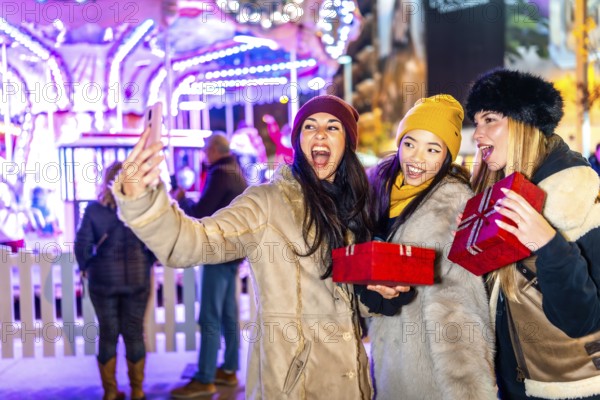 Three cheerful women holding christmas gifts taking a selfie in front of a carousel at night in a christmas themed amusement park