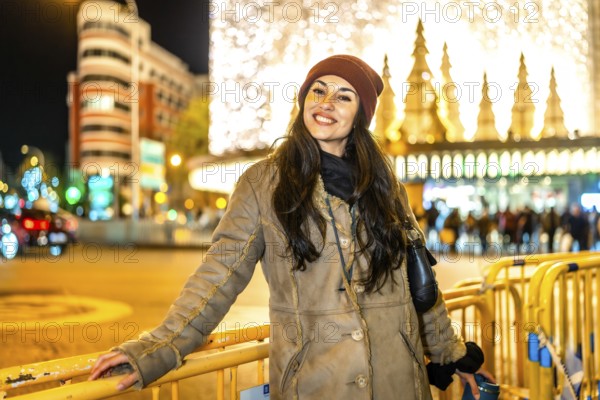 Smiling young woman leaning on a fence in a city street enjoying christmas lights and decorations at night
