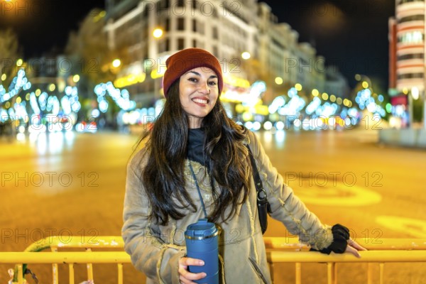 Young woman wearing winter clothes, holding a thermal mug and smiling near a street fence with christmas lights in the background