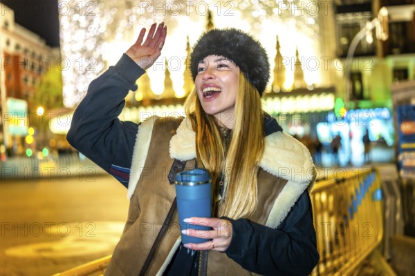 Young woman greeting someone while enjoying a hot beverage in a reusable cup in a city decorated with christmas lights