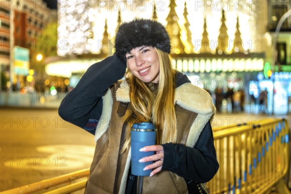 Stylish woman holding reusable cup with hot drink, enjoying magical christmas lights and festive atmosphere in a city at night