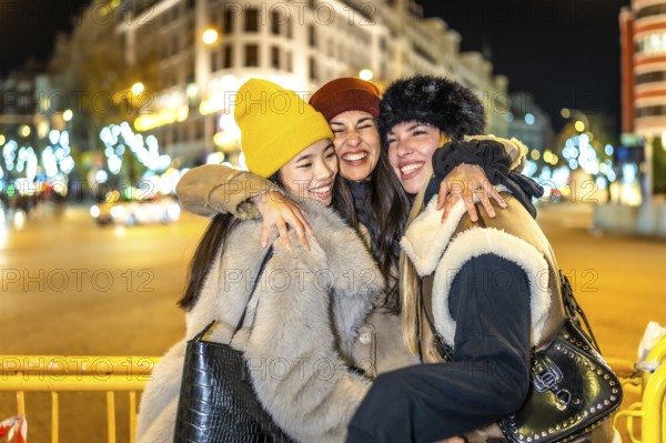 Three cheerful young women hugging each other in a city street decorated with christmas lights