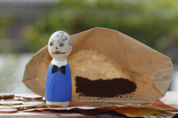 Male wooden cone figure with coins and banknote in front of filter bag with coffee North Rhine-Westphalia, Germany