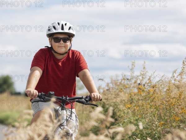 A boy with a bicycle helmet rides a bicycle and smiles friendly into the camera, AI generated, AI generated