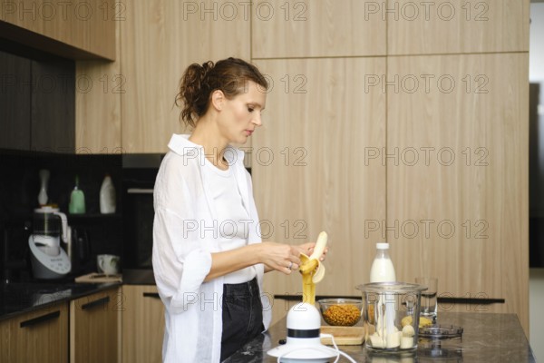 In a stylish kitchen, a woman is focused on preparing a smoothie by peeling a banana. Nearby, a glass container holds sea buckthorn berries and a bottle of milk