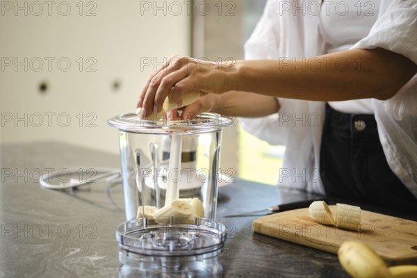 Sliced bananas are placed in a blender while a sunny kitchen provides a bright backdrop. Fresh ingredients are used for a nutritious sea buckthorn smoothie. The atmosphere is vibrant and inviting