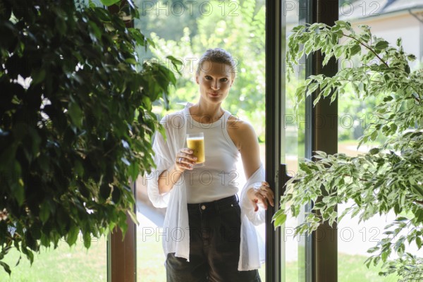 A woman stands in a doorway holding a glass of refreshing beverage. Sunlight filters through greenery, creating a warm atmosphere. She appears relaxed, enjoying the pleasant afternoon