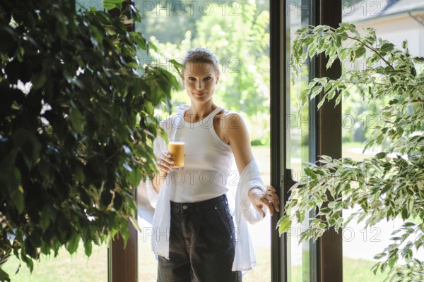 A young woman stands at the entrance of a bright garden space, holding a glass of iced beverage. Dressed in a casual white top and jeans, she appears relaxed amid lush green plants in the background