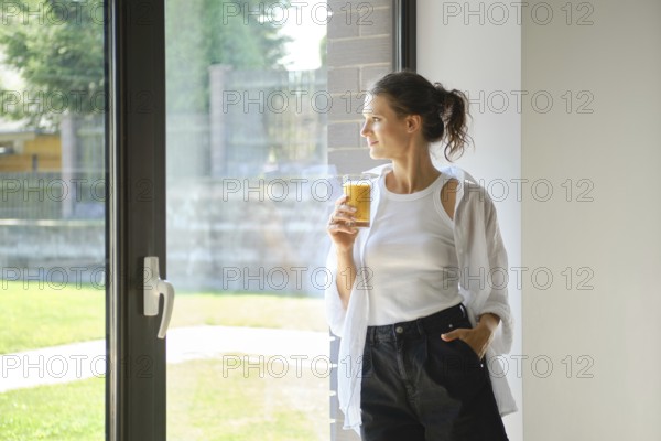 A woman in a white top and black pants stands by a window, holding a glass of smoothies. She gazes outside, enjoying the warm sunlight and the serene view of greenery