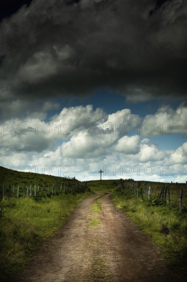 A winding dirt road leads the eye to a solitary cross. Cézallier, Auvergne Volcanoes Regional Natural Park, Puy-de-Dôme, Auvergne Rhone Alpes, France