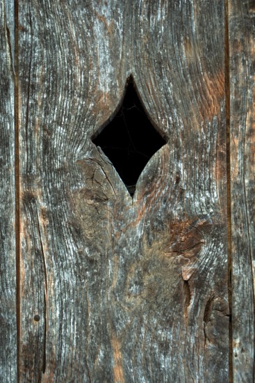 A close-up view of a weathered wooden panel, featuring a diamond-shaped opening cut into the surface. The wood displays signs of age with a rich texture and grain
