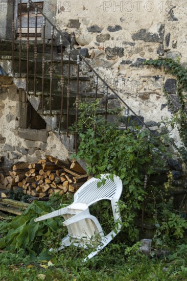 An unkempt outdoor area features a white chair leaning against a weathered stone wall, surrounded by creeping vines and overgrown plants. Nearby, a stack of firewood illustrates neglect. France