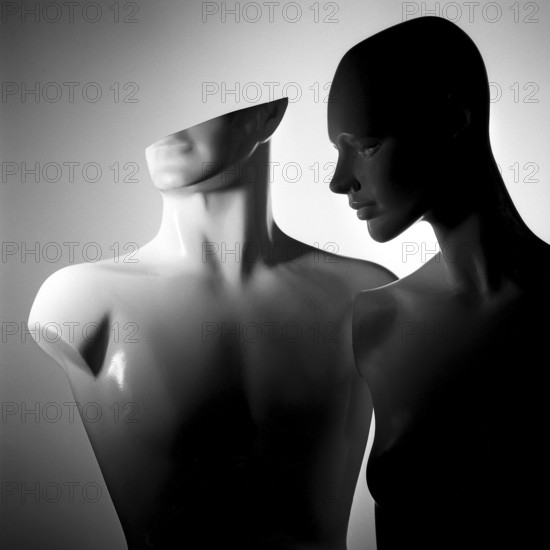 Two stylized mannequins stand closely together, featuring smooth surfaces and abstract shapes. Artistic lighting creates a captivating interplay of light and shadow, highlighting their distinct forms