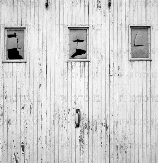 The weathered wooden wall, painted white, showcases three cracked windows that reveal a sense of age and history. This rustic structure captures a timeless, vintage aesthetic