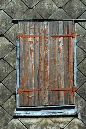 Closed wooden shutters with peeling paint rest against an aged building, highlighting the rustic charm of its textured roof tiles under soft natural light. France