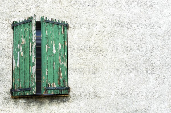 Weathered green shutters stand slightly ajar on a textured white wall, showcasing the charm of an old building and reflecting years of history and character
