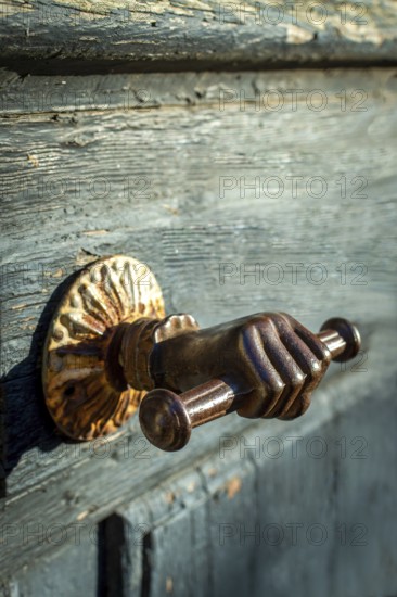 A distinctive door handle resembling a hand grips a rustic wooden door, featuring intricate details and a charming patina. The warm light enhances the texture and design